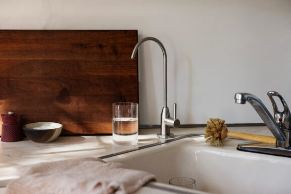 A kitchen sink with a glass of water, a dish brush, a wooden cutting board, a towel, and a small bowl on the counter in natural light.