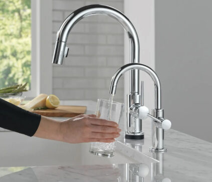 A hand holds a glass under a running faucet on a marble countertop next to another chrome faucet in a modern kitchen, where sleek appliances and massage guns hint at a lifestyle focused on wellness and convenience.