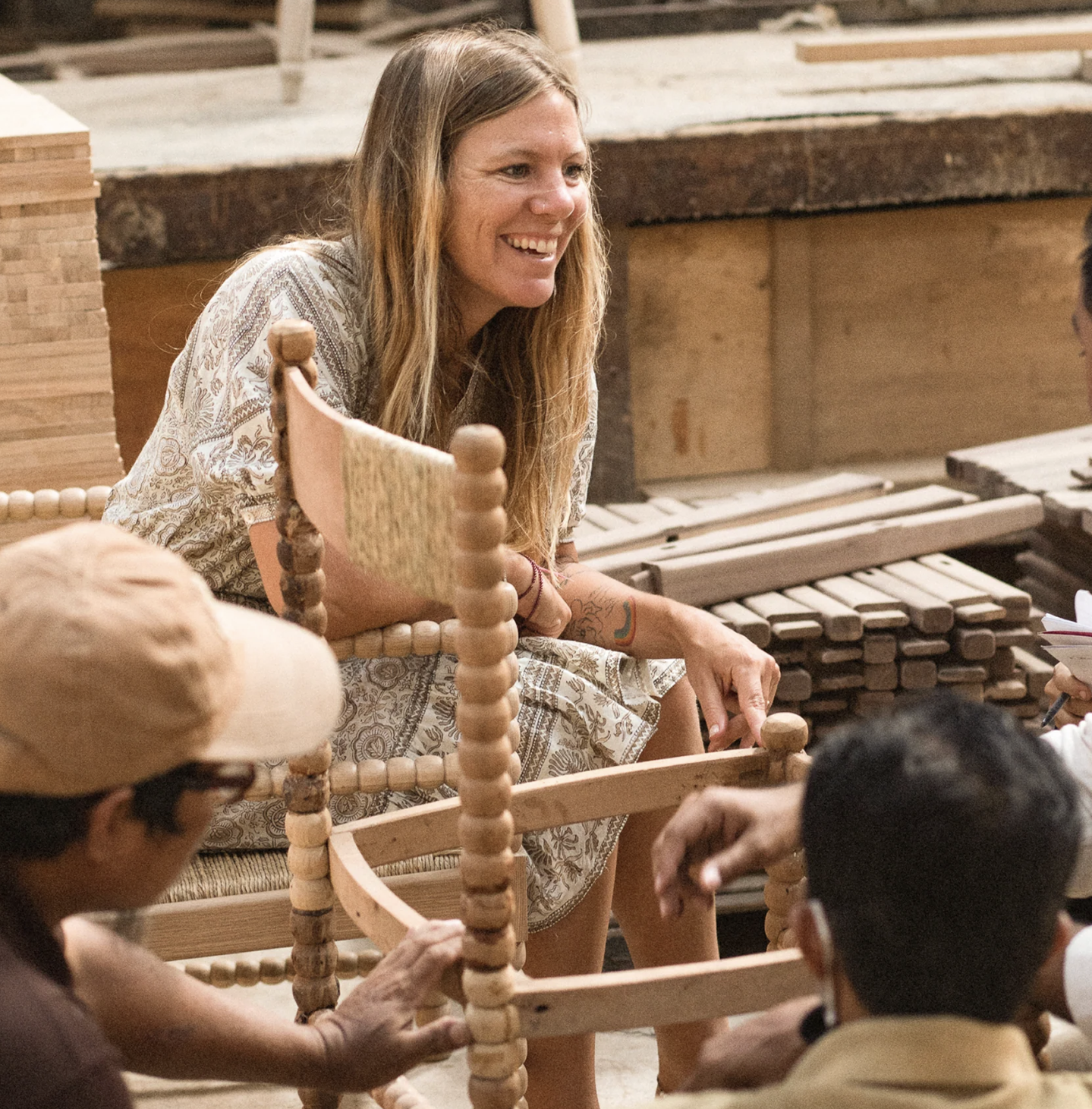 A woman sits and smiles while talking to three people in a workshop, surrounded by wooden planks and a partially assembled wooden chair.