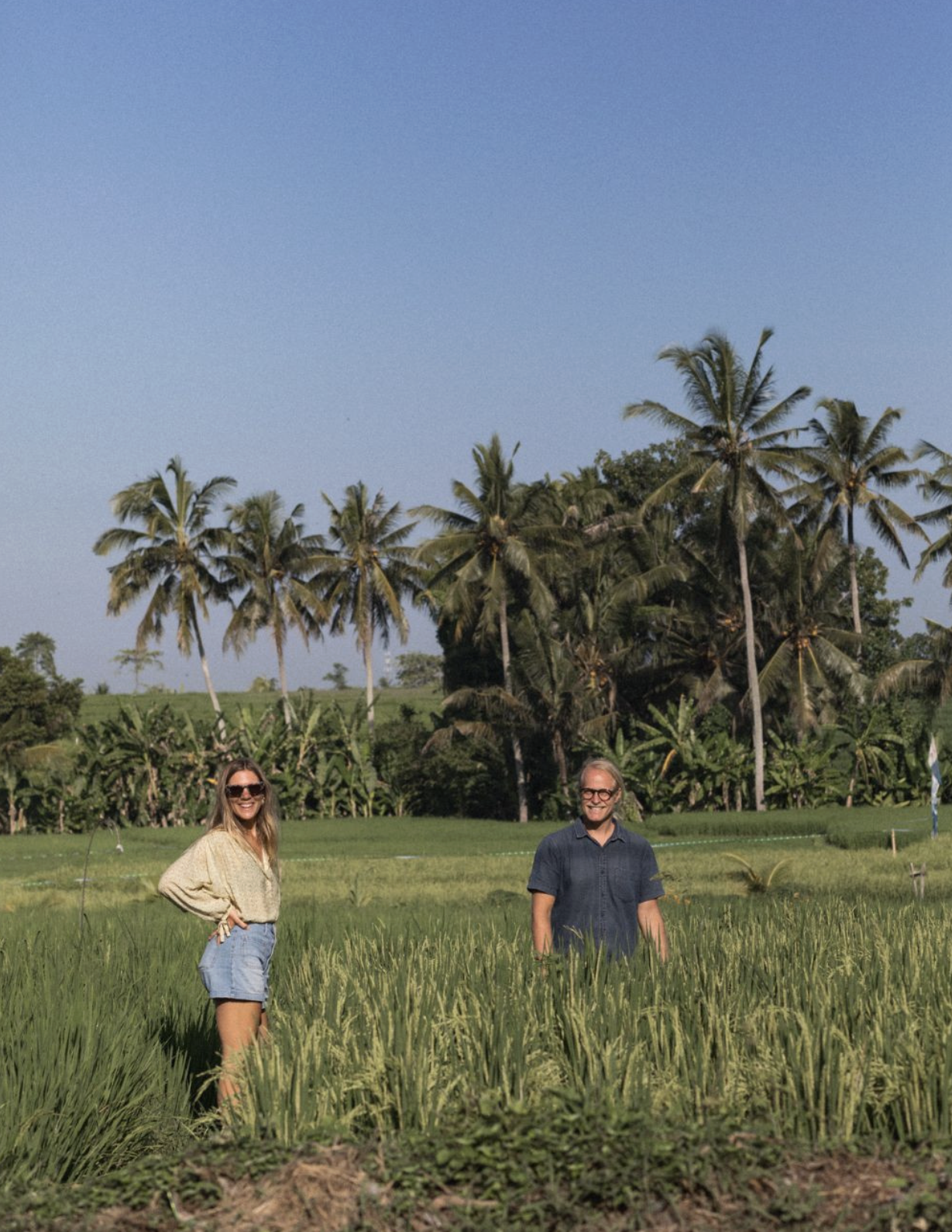 Two people stand in a green field with tall grass, palm trees, and clear blue sky in the background.