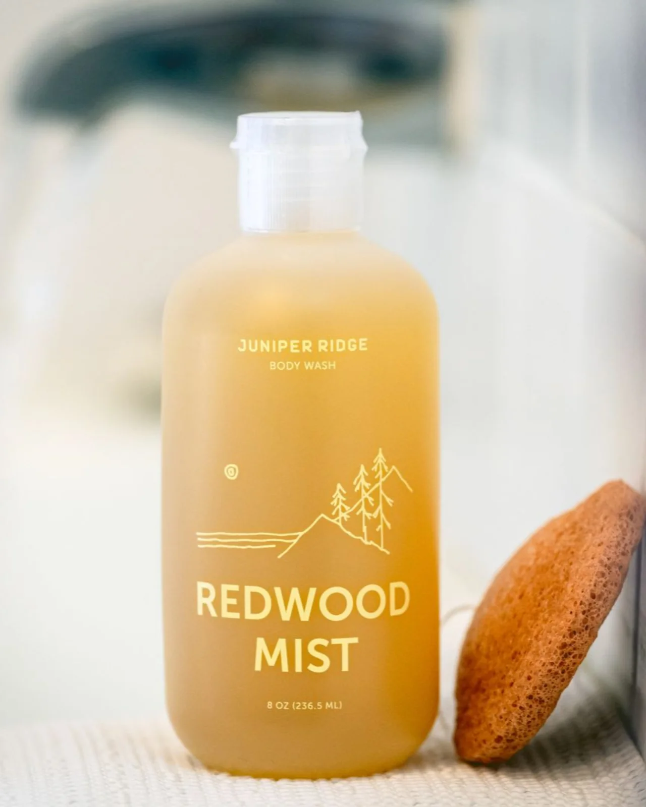 A bottle of Juniper Ridge Redwood Mist body wash stands next to a tan bath sponge on a bathroom surface.