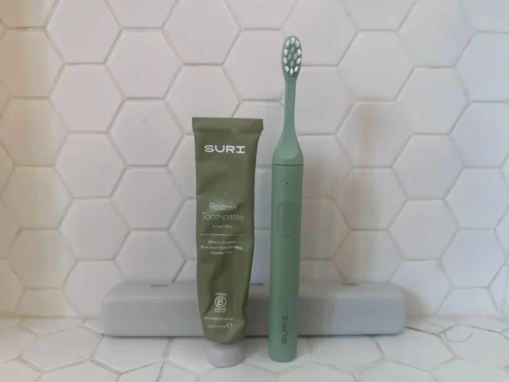 Green SURI toothpaste tube and matching green electric toothbrush standing upright on a white hexagonal tile surface, with two gray toothbrush cases in the background.