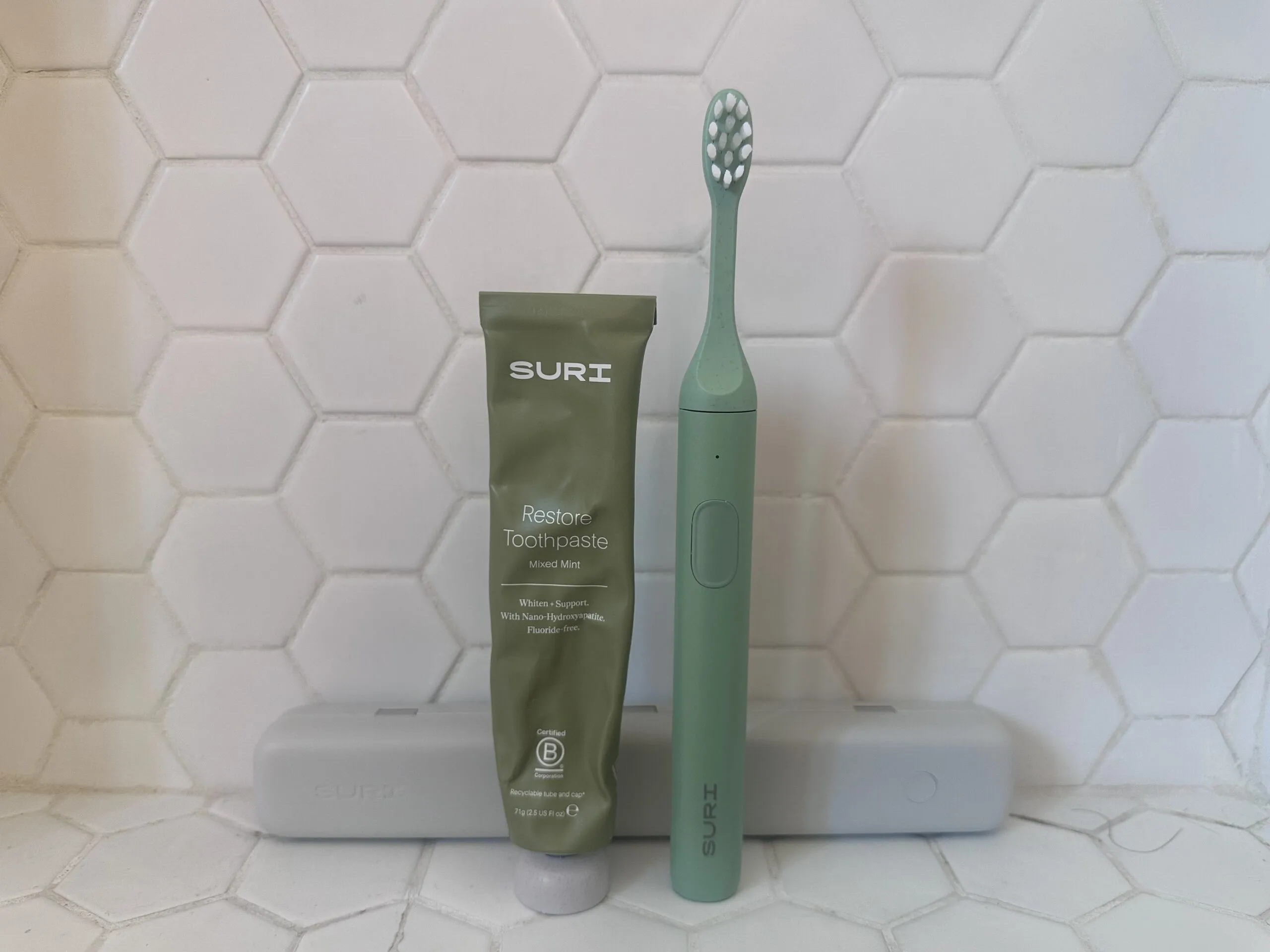 Green SURI toothpaste tube and matching green electric toothbrush standing upright on a white hexagonal tile surface, with two gray toothbrush cases in the background.