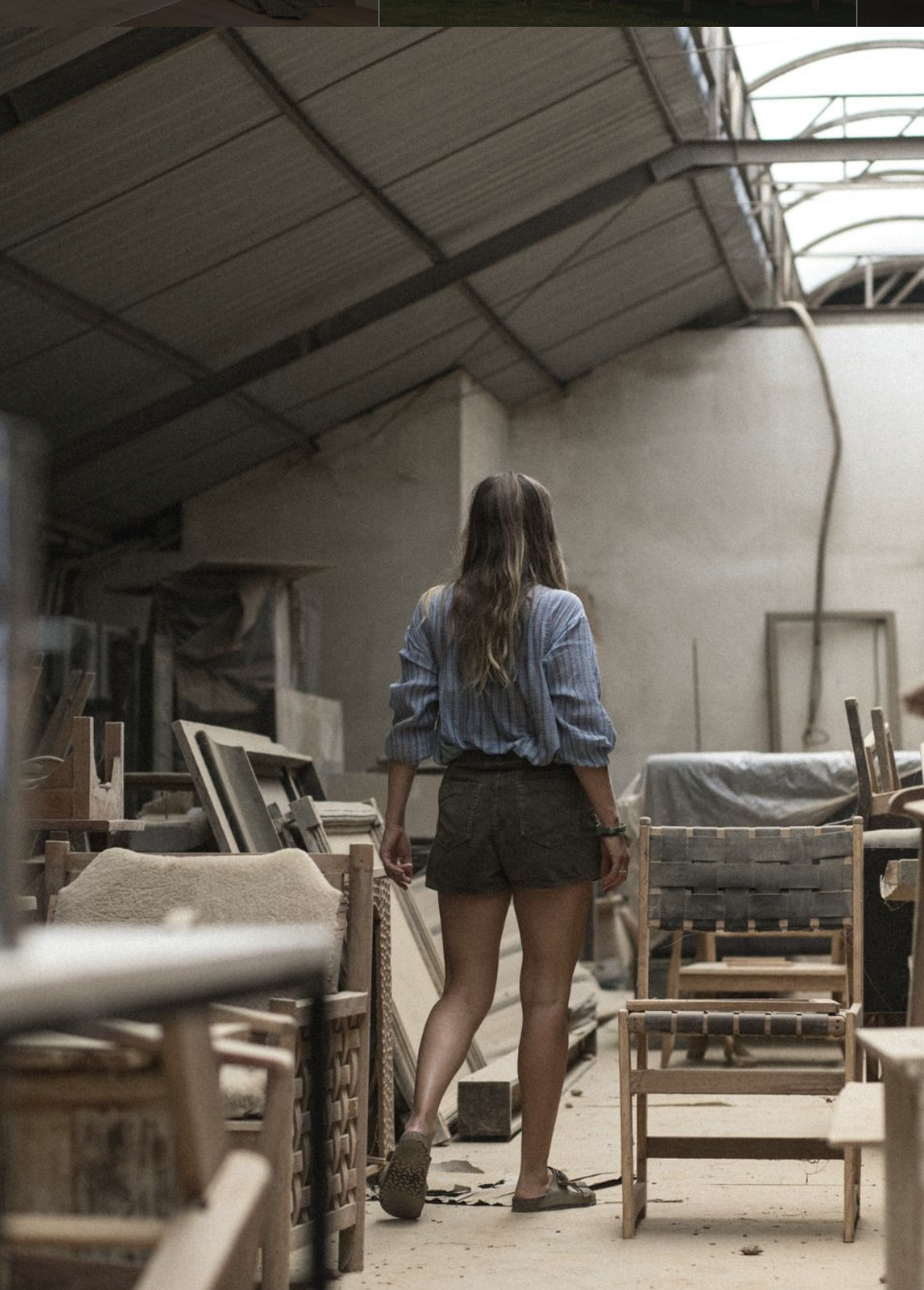 A woman with long hair, wearing a blue shirt and shorts, walks through a cluttered workshop filled with wooden furniture and tools.