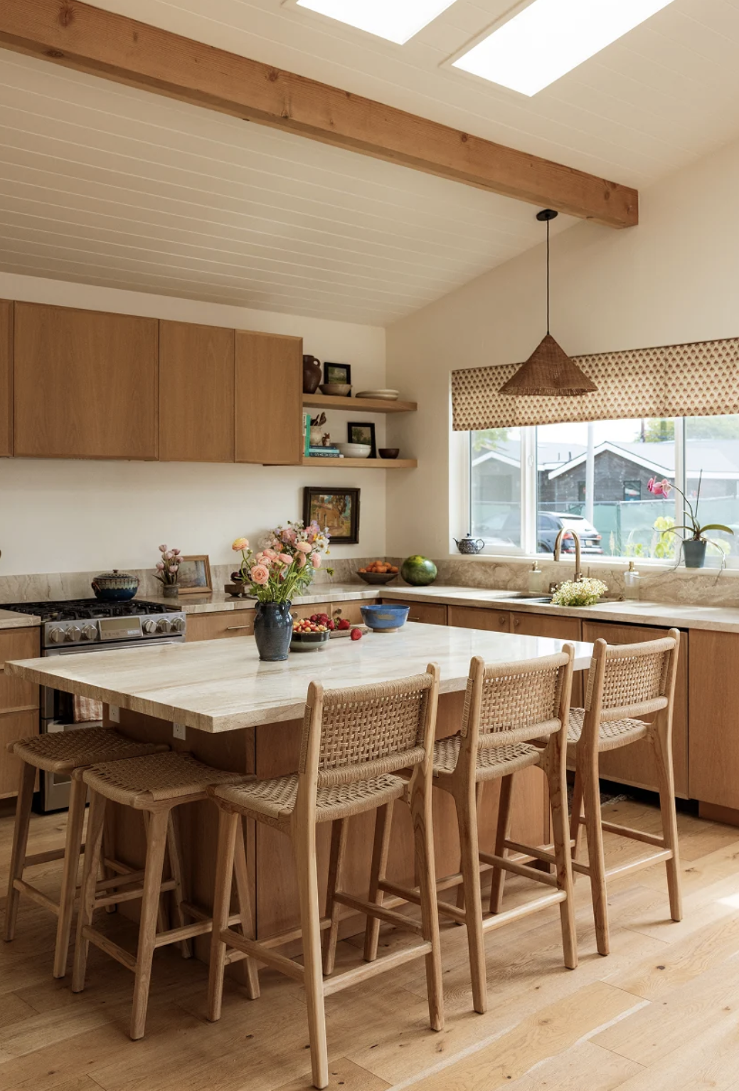 A modern kitchen with wooden cabinets, a marble island, four woven bar stools, flowers in a vase, and sunlight coming through a window and skylights.