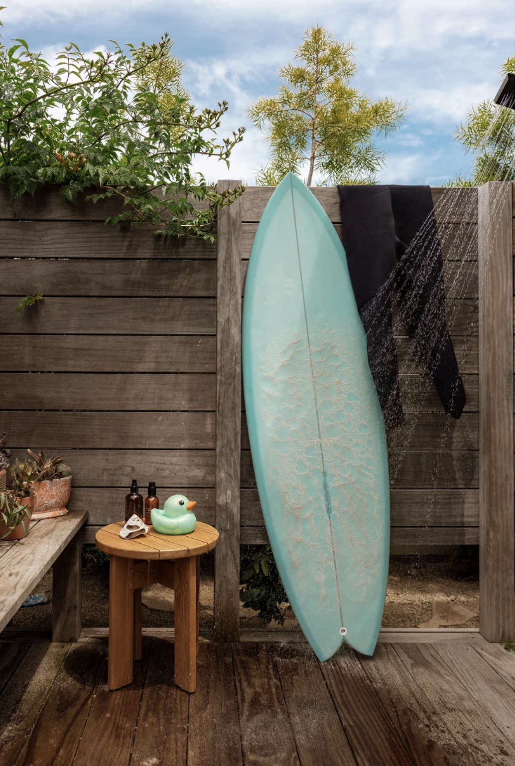 A blue surfboard leans against an outdoor wooden shower wall with a black wetsuit, next to a small table holding bottles and a green duck.