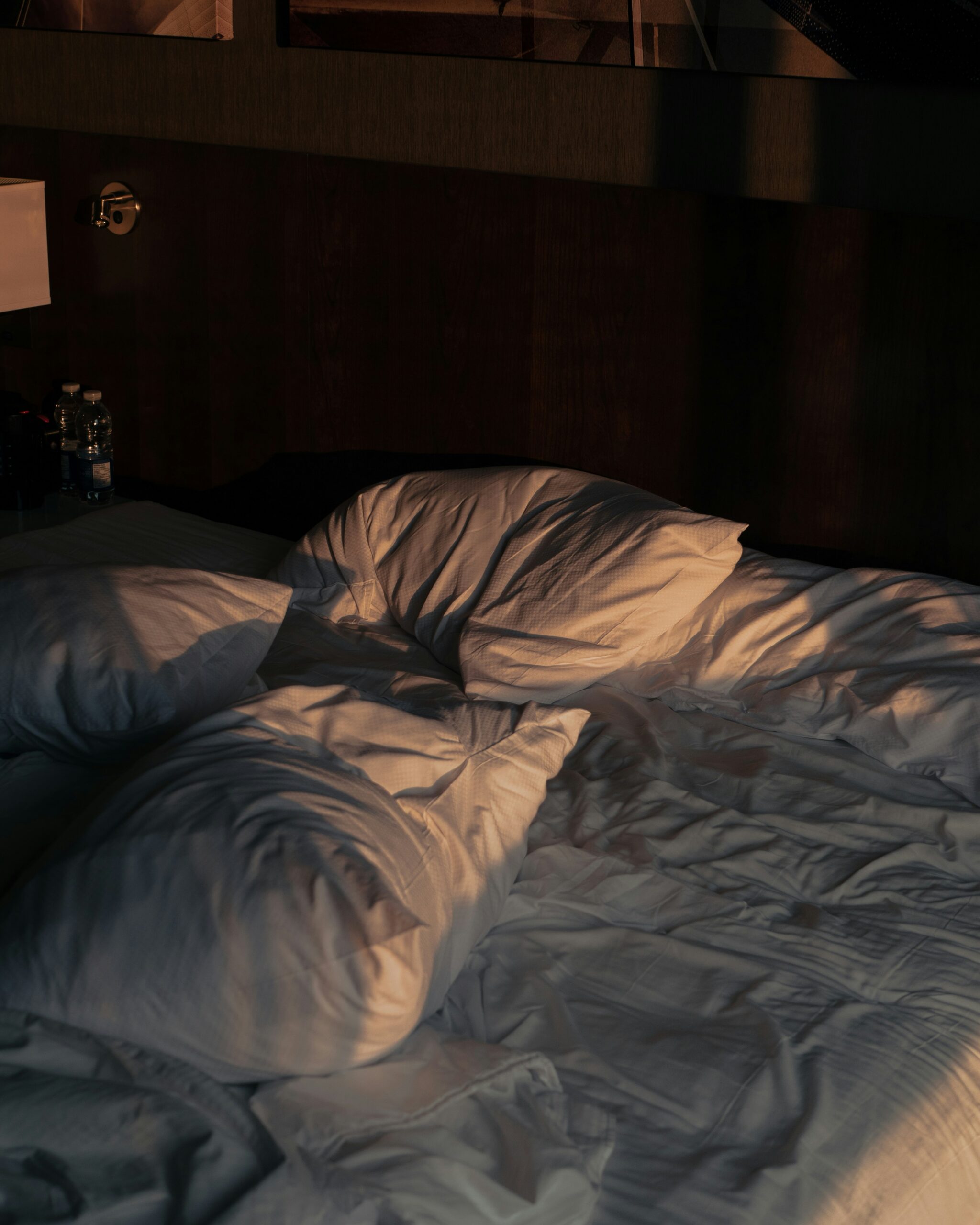 Unmade bed with white pillows and sheets in dim lighting. Two water bottles are visible on a nightstand beside the bed.