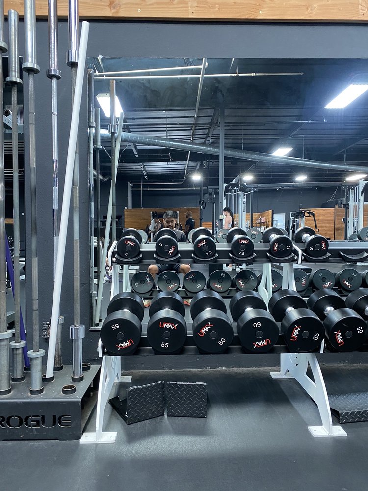 Rows of dumbbells on racks in front of a large mirror at a gym, with people working out and gym equipment reflected in the background.