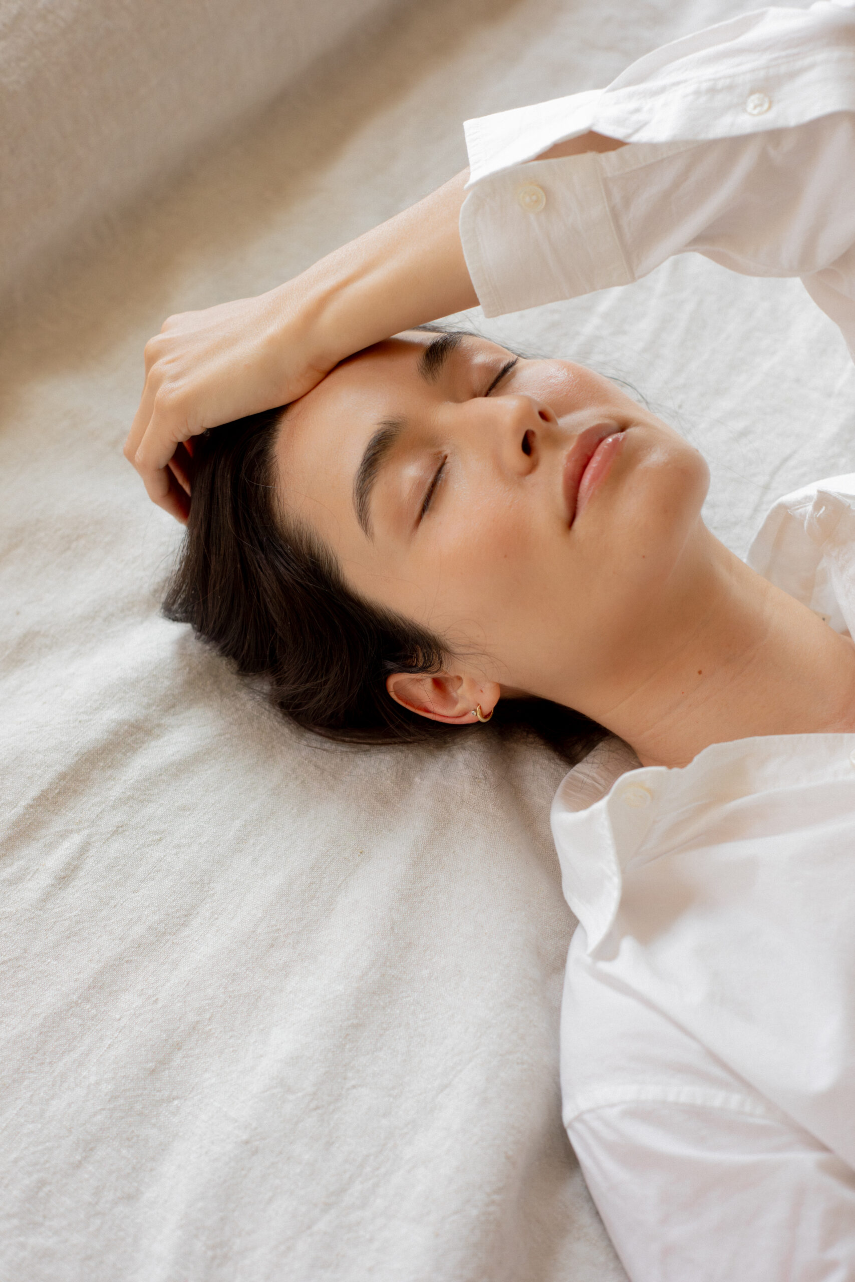 A person in a white shirt lies on a light-colored bed with eyes closed and one hand resting on their forehead.