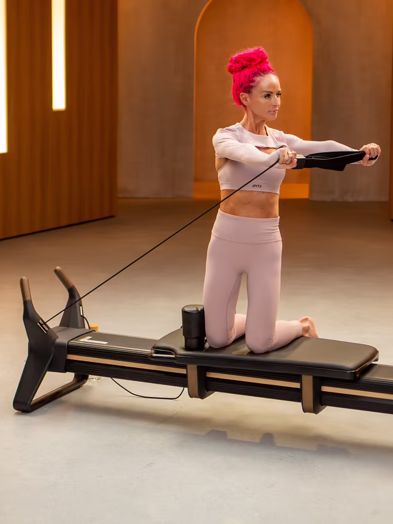 Person with pink hair kneeling on a home Pilates reformer, pulling resistance cords in a modern studio with wooden panel walls and vertical lights.