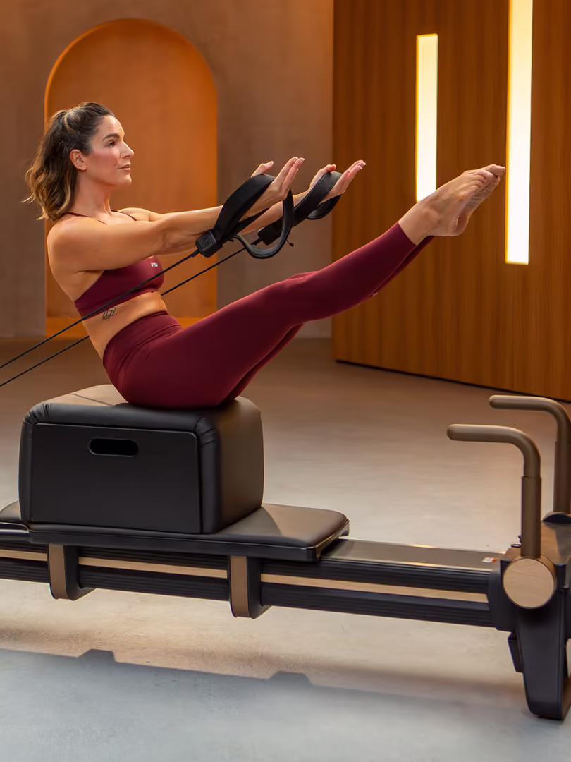 A person in workout attire sits on a Pilates reformer, holding straps and lifting their legs, performing an exercise in a modern, well-lit studio equipped with home Pilates reformers.