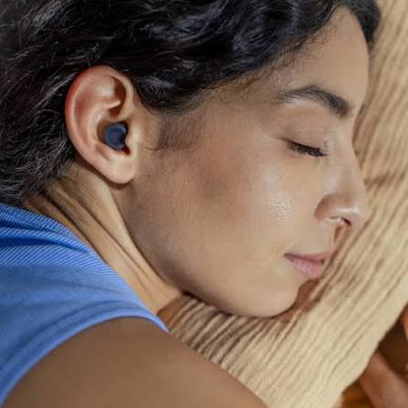 A person with dark hair sleeps on their side with a wireless earbud in their ear, resting their head on a beige pillow.