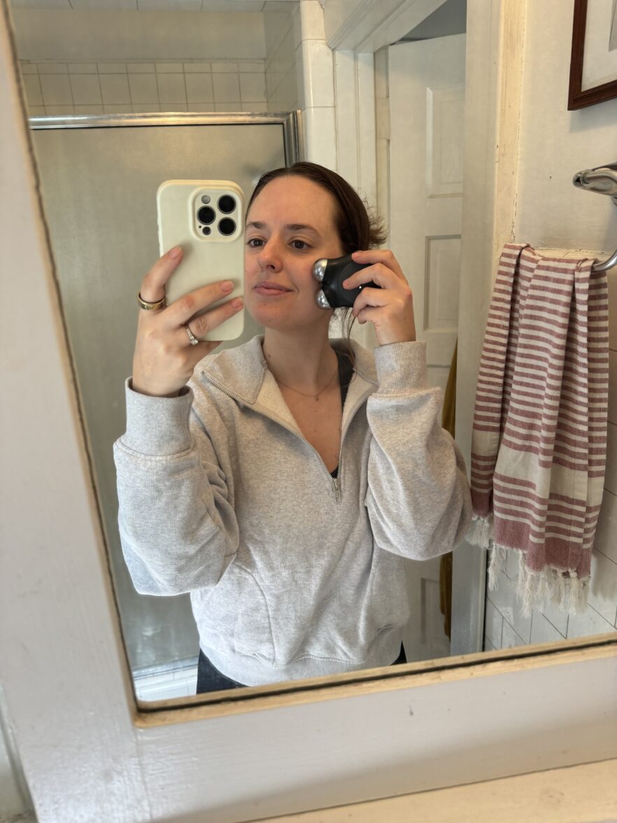 Person stands in a bathroom, looking into a mirror while holding an electric shaver to their cheek, wearing a light gray zip-up sweatshirt.