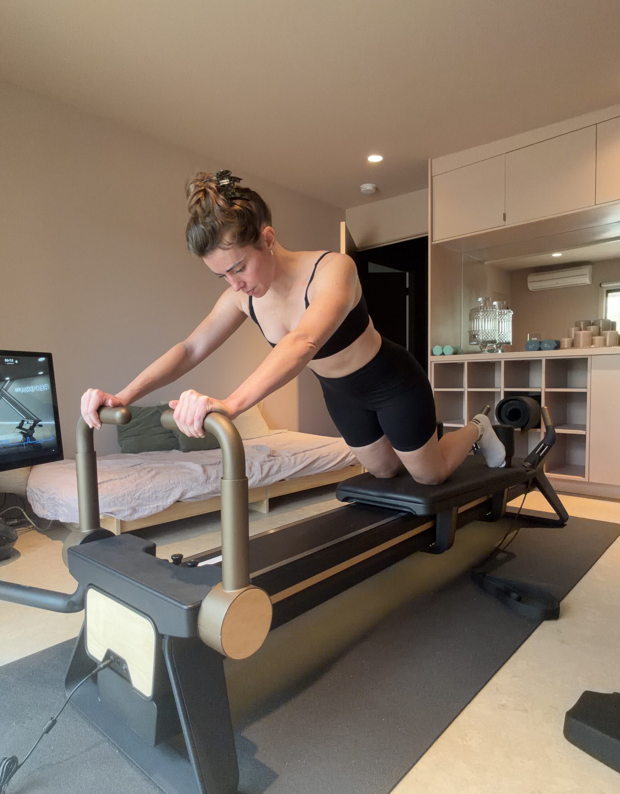 A woman performing a pilates exercise on a reformer machine in a home room.