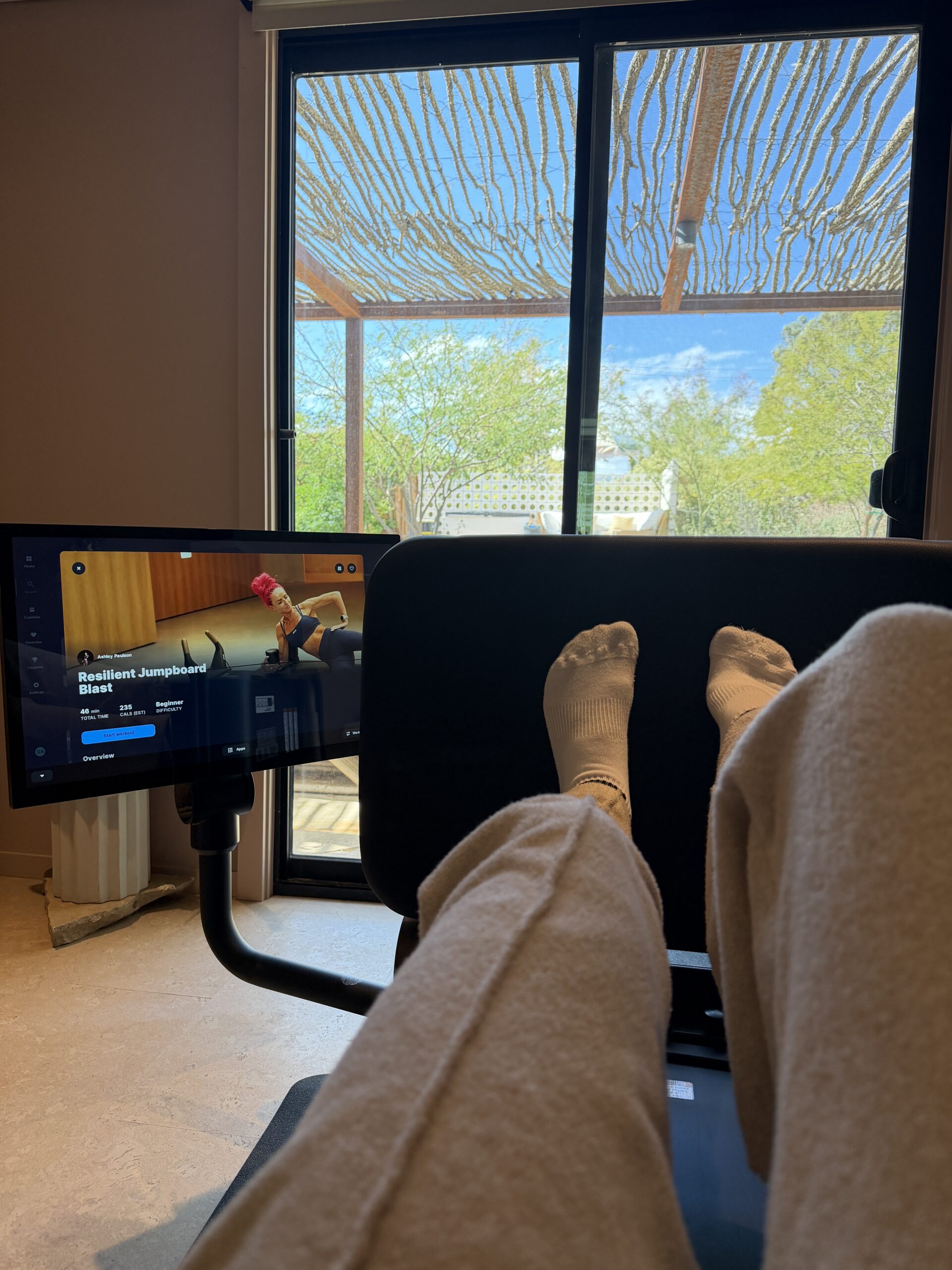 A person in sweatpants and white socks on a Pilates reformer, facing a screen showing a fitness instructor performing a jumpboard workout.