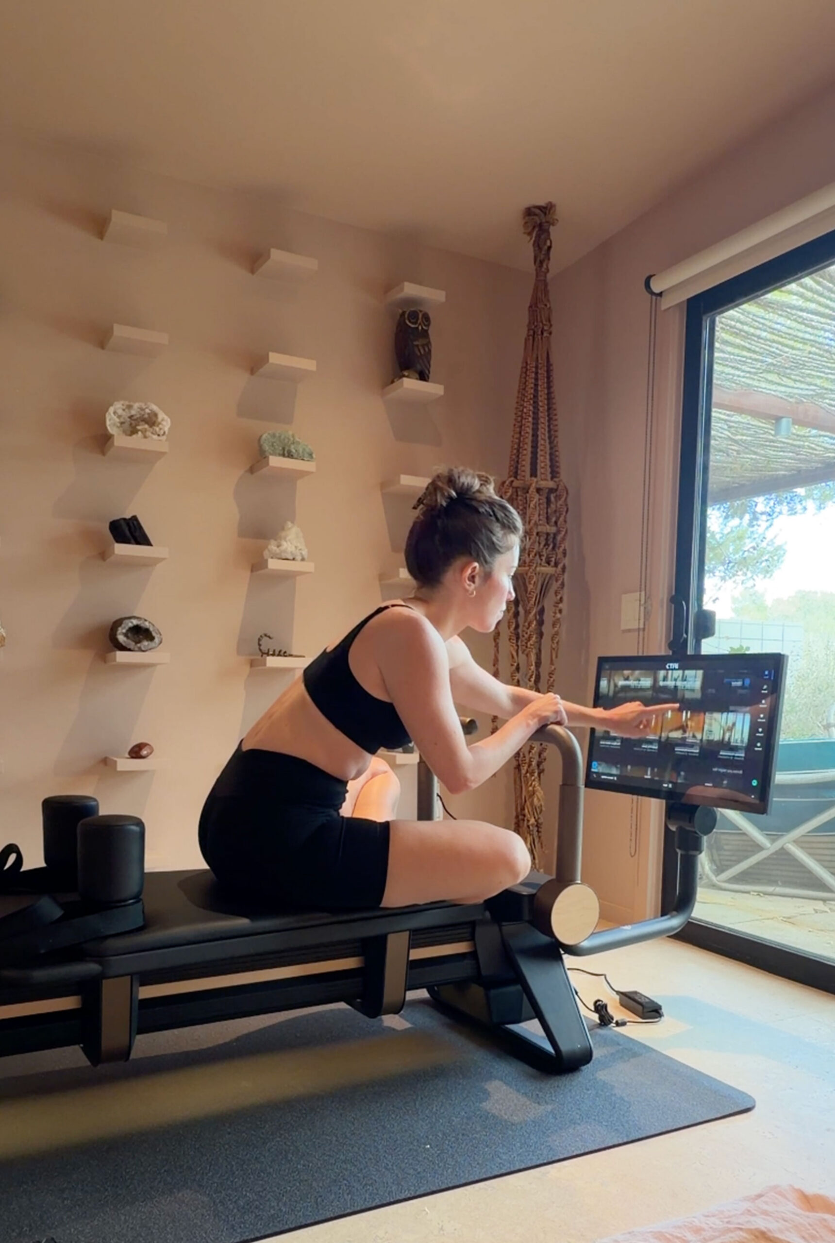 A woman in workout clothes sits on a Pilates reformer and touches the screen of an attached tablet.
