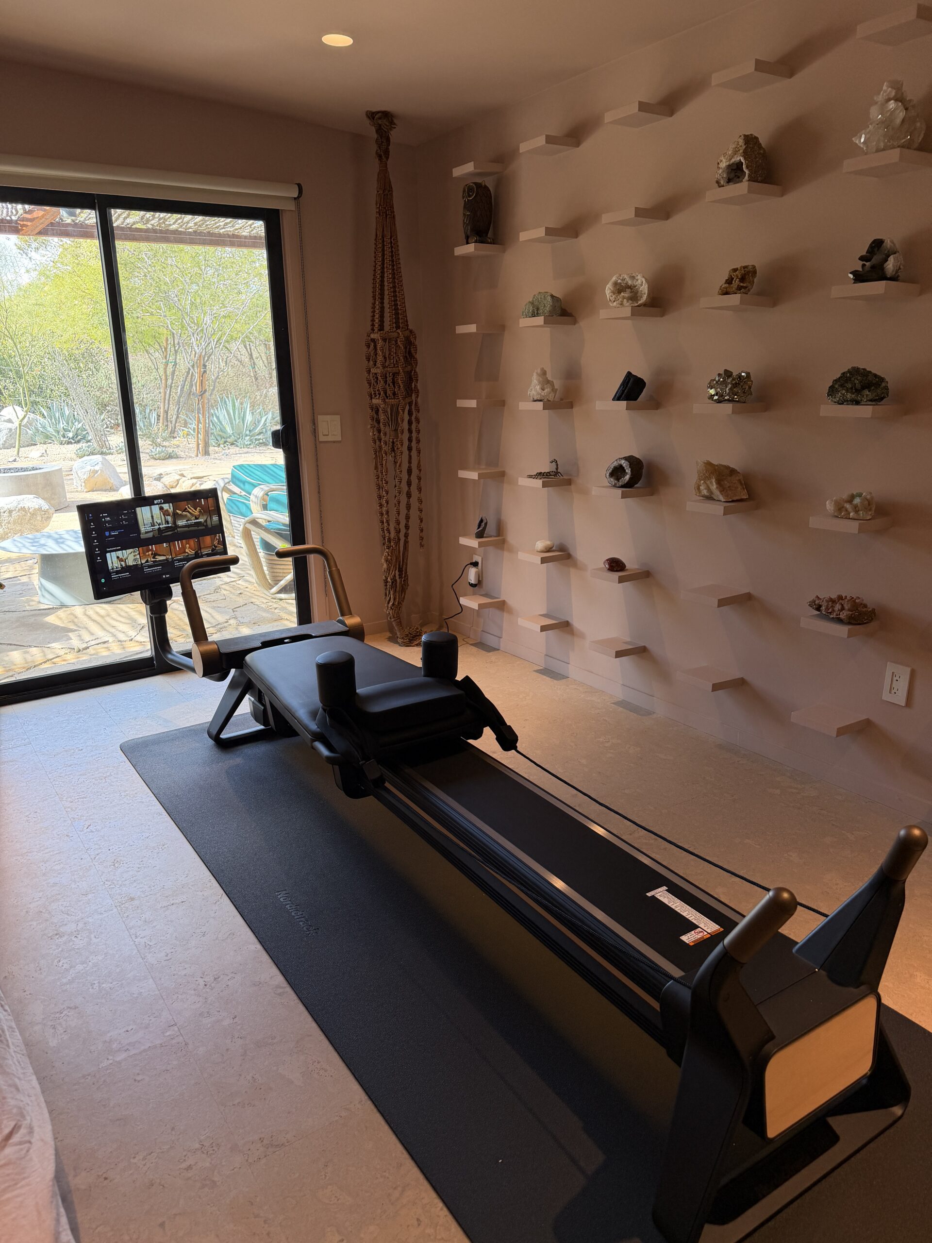 A Pilates reformer machine on a black exercise mat in a room with shelves displaying various rocks and crystals.