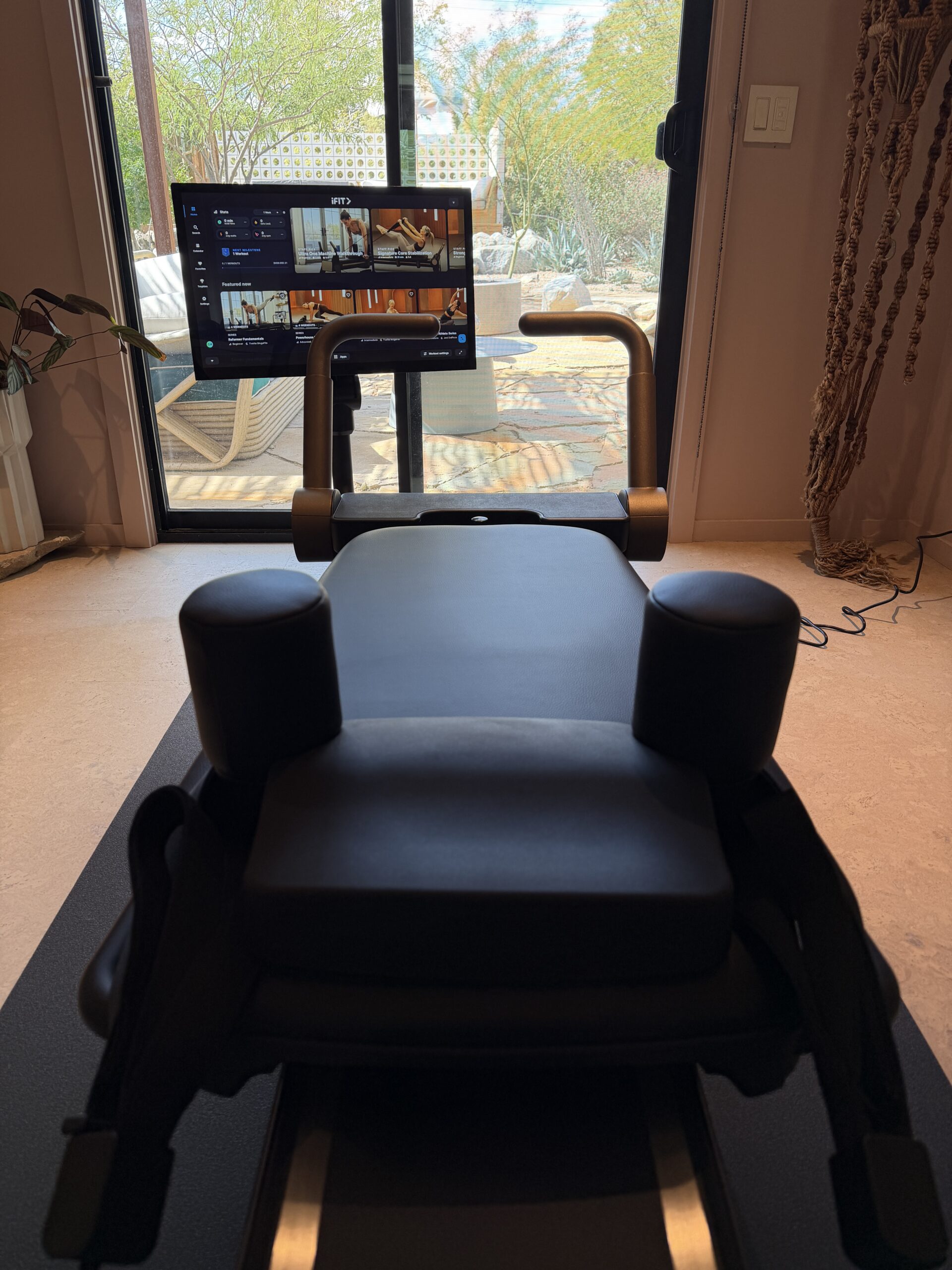 A Pilates reformer machine positioned in front of a glass door, with a screen displaying an iFIT workout interface.