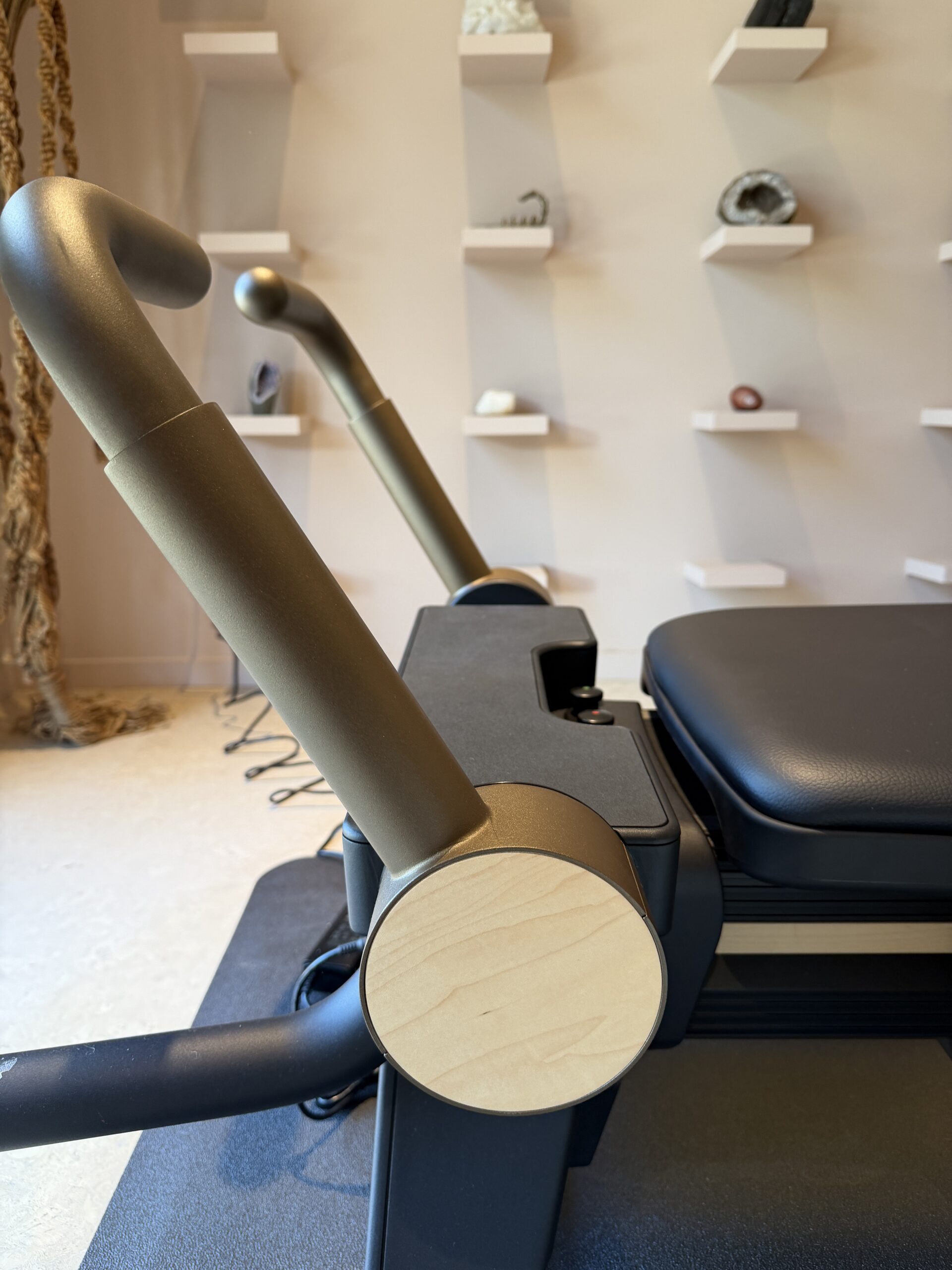 A Pilates reformer machine with matte gold upright handles and a black padded carriage, positioned in front of a wall with small display shelves.