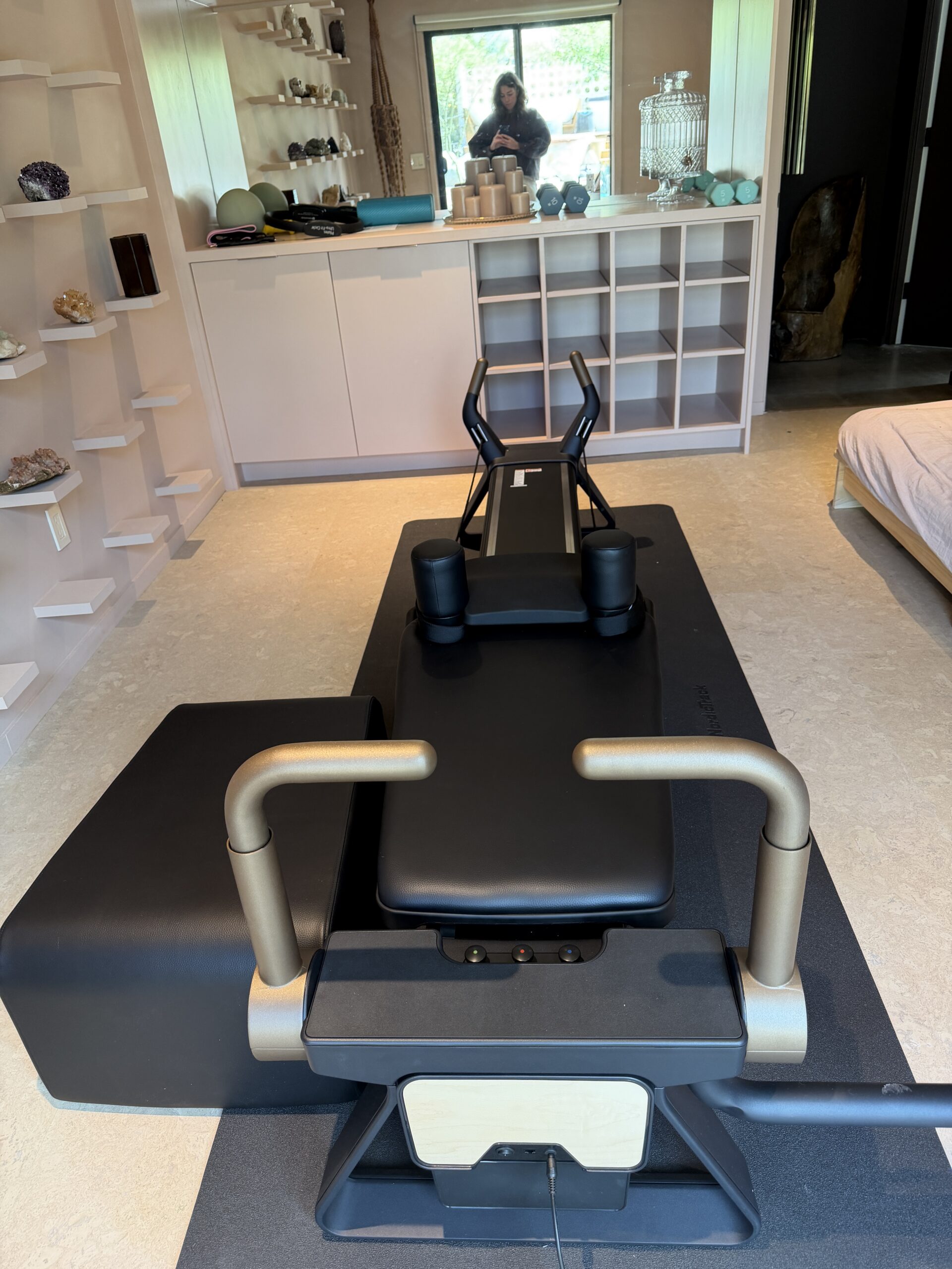 A black Pilates reformer machine positioned on a mat in a room with built-in shelving.